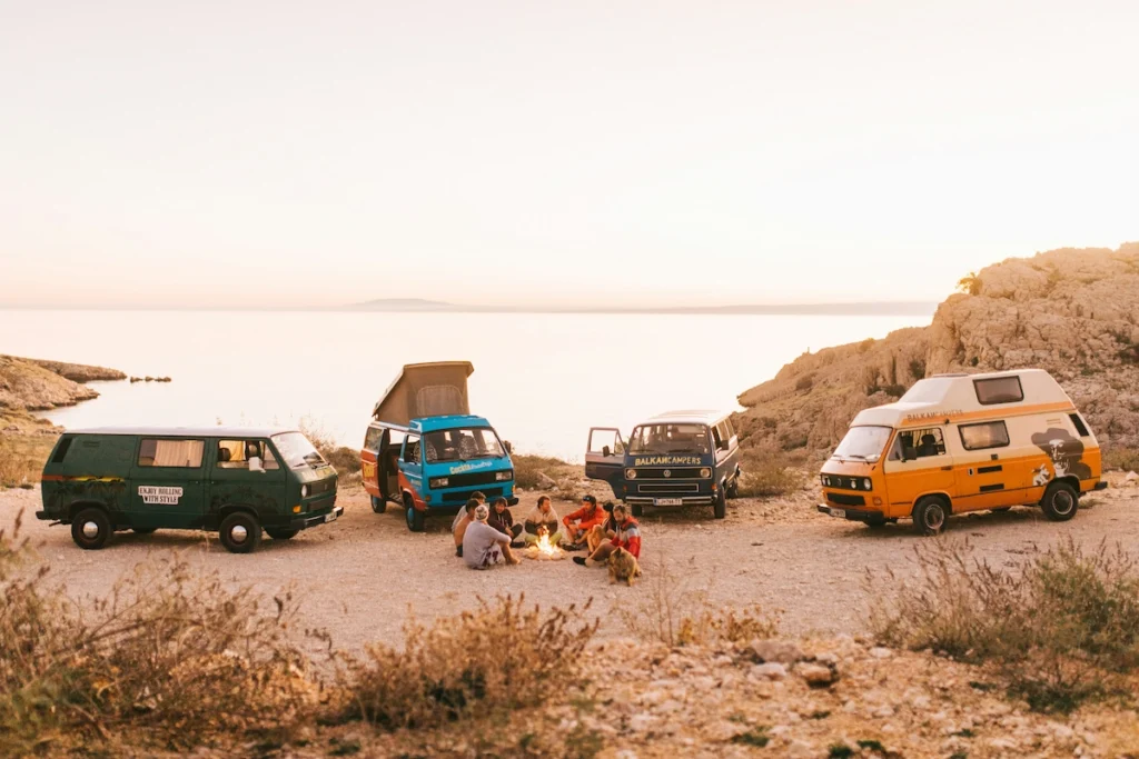 A picture of a small gathering of a small group of van lifers. They are all sitting beside a camp fire and their camper vans are parked behind.