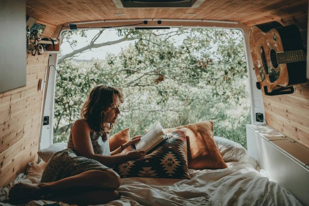 An woman reeding a book on the bed inside her van parked in nature. The opene
