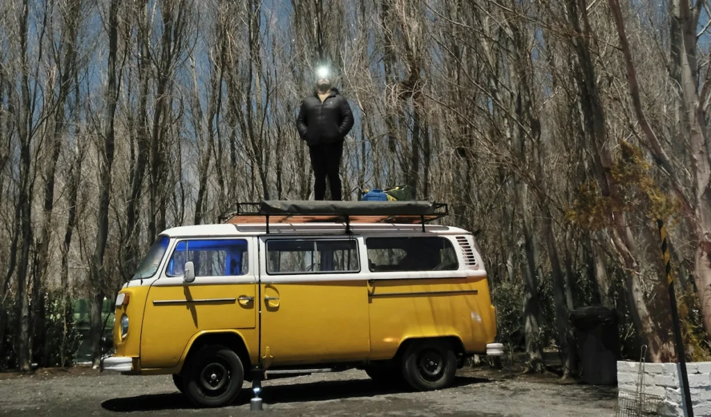 A man standing on top of camper van checking the surroundings with a light. The van is parked on a dispersed land.  