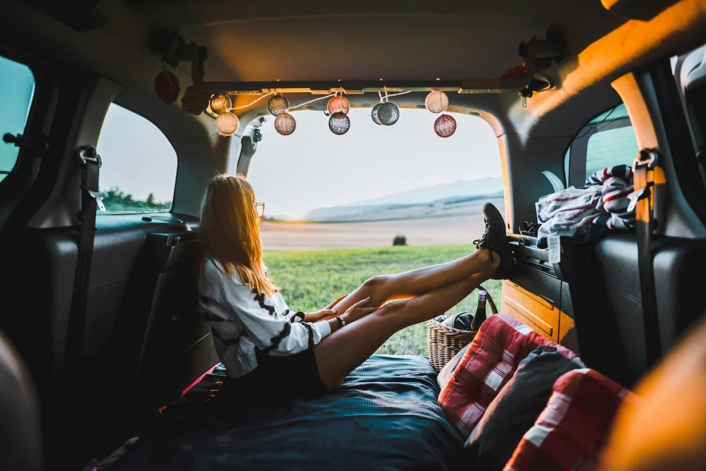 A young van lifer girl sitting inside her camper van with minimal and simple set-up of a bed and storage.
